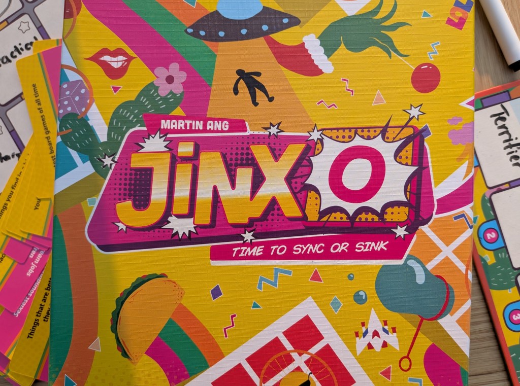 A close up of the JinxO box with some components (player boards, markers) in the background.